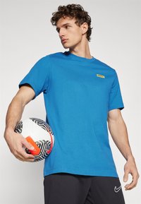 Camiseta de algodón azul de manga corta con cuello redondo, que presenta un logo amarillo de Nike FC en el pecho. Se sostiene un balón de fútbol bajo el brazo.