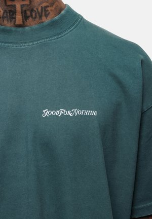 Henkilö, jolla on päällään teal-värinen paita, jonka rinnassa on valkoisella tekstillä "HYÖDYTTÖMÄ" ja kaulassa näkyvä tatuointi, jossa lukee "PELÄÄ RAKASTAA".