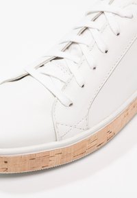 Vit lädersko med vita snören, med en texturerad korksula och minimala designelement, som framhäver rena linjer och släta ytor.