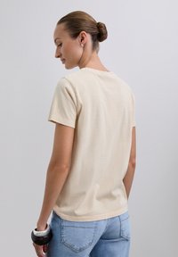 Camiseta de algodón beige con cuello redondo y mangas cortas, que presenta un corte holgado y una textura suave, combinada con vaqueros azules.