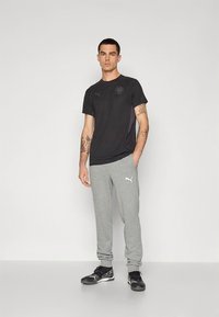 Juoda Puma marškinėliai su logotipu, derinami su šviesiai pilkomis sportinėmis kelnėmis. Modelis dėvi juodus sportinius batus. Paprastas, sportiškas dizainas ir audinio tekstūra.