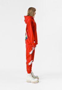 Sudadera roja con estampado gráfico, combinada con pantalones de chándal rojos que presentan diseños de plumas blancas; llevada con botas beige de cordones.