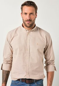 Jasnobrązowa koszula zapinana na guziki z małym haftowanym wzorem, z długimi rękawami podwiniętymi na mankietach, w połączeniu z niebieskimi dżinsami i brązowym paskiem.