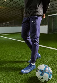 Modré futbalové kopačky s textúrovanými detailmi, v kombinácii s námorníckymi športovými nohavicami s bielym logom, na umelom trávniku vedľa vzorovanej futbalovej lopty.