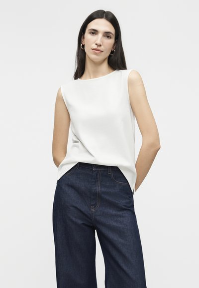 Γυναίκα με μακριά σκούρα μαλλιά, φοράει χρυσούς κρίκους σκουλαρίκια, λευκό αμάνικο τοπ και σκούρα μπλε τζιν με ψηλή μέση και φαρδιά πόδια, χέρια πίσω από την πλάτη.