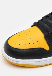 Svart och gul sneaker med övre i slätt läder, perforerat mönster på tån och texturerad gummisula, med svarta snören.