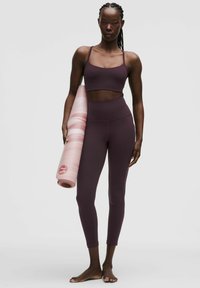 Frau in lila Sport-BH und Leggings, die eine gerollte rosa Yogamatte hält und barfuß vor einem schlichten grauen Hintergrund steht.