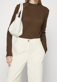 Bruine longsleeve top met een hoge halslijn, gecombineerd met crèmekleurige wijde broek. Een witte leren handtas hangt over de arm.