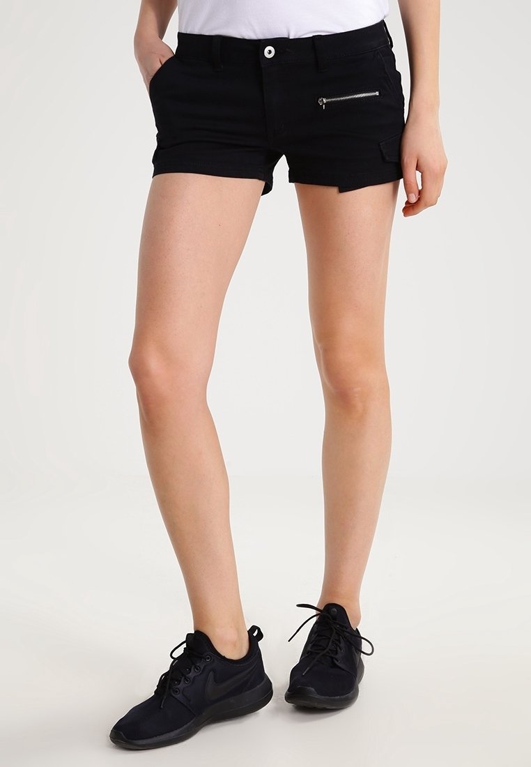 Femme portant un short noir à poches zippées, un haut blanc et des baskets noires Nike, debout les mains dans les poches sur fond blanc.