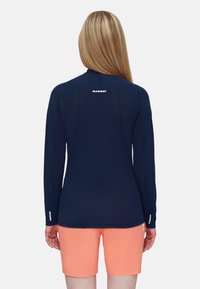 Marineblaues Langarmhemd mit hohem Kragen und hellem Stickmuster, kombiniert mit korallenfarbenen Shorts. Auf der Rückseite befindet sich ein weißes Logo.