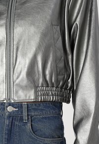 Metalni srebrni bomber jakna z zadrgo spredaj, rebrastim robom in teksturiranimi detajli; v kombinaciji z visoko pasom modrimi denim hlačami.