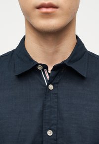 Zbliżenie na szyję i podbródek w granatowej koszuli na guziki z białą i czerwoną paskowaną lamówką wewnątrz kołnierzyka.