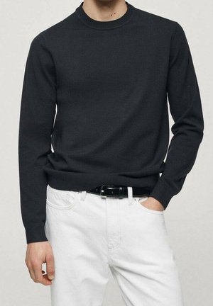 Homme portant un pull noir ajusté à manches longues rentré dans un pantalon blanc avec une ceinture noire, une main dans la poche.