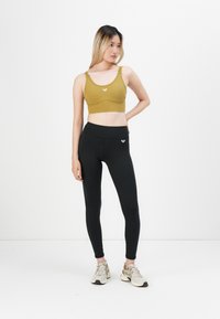 Woman wearing olive sports bra, black leggings, and beige sneakers standing against white background with one hand on hip.