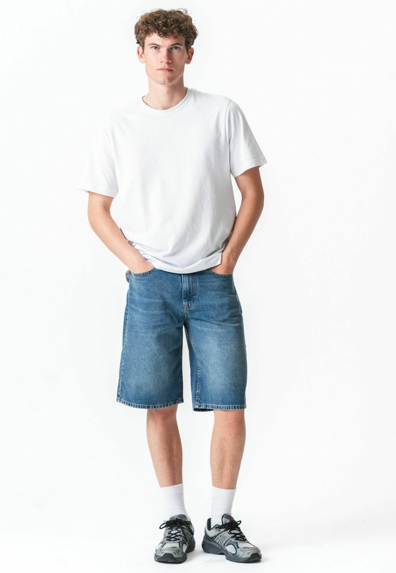 Jeune homme debout les mains dans les poches, portant un t-shirt blanc, un short en jean jusqu'aux genoux, des chaussettes blanches et des baskets grises sur fond blanc.