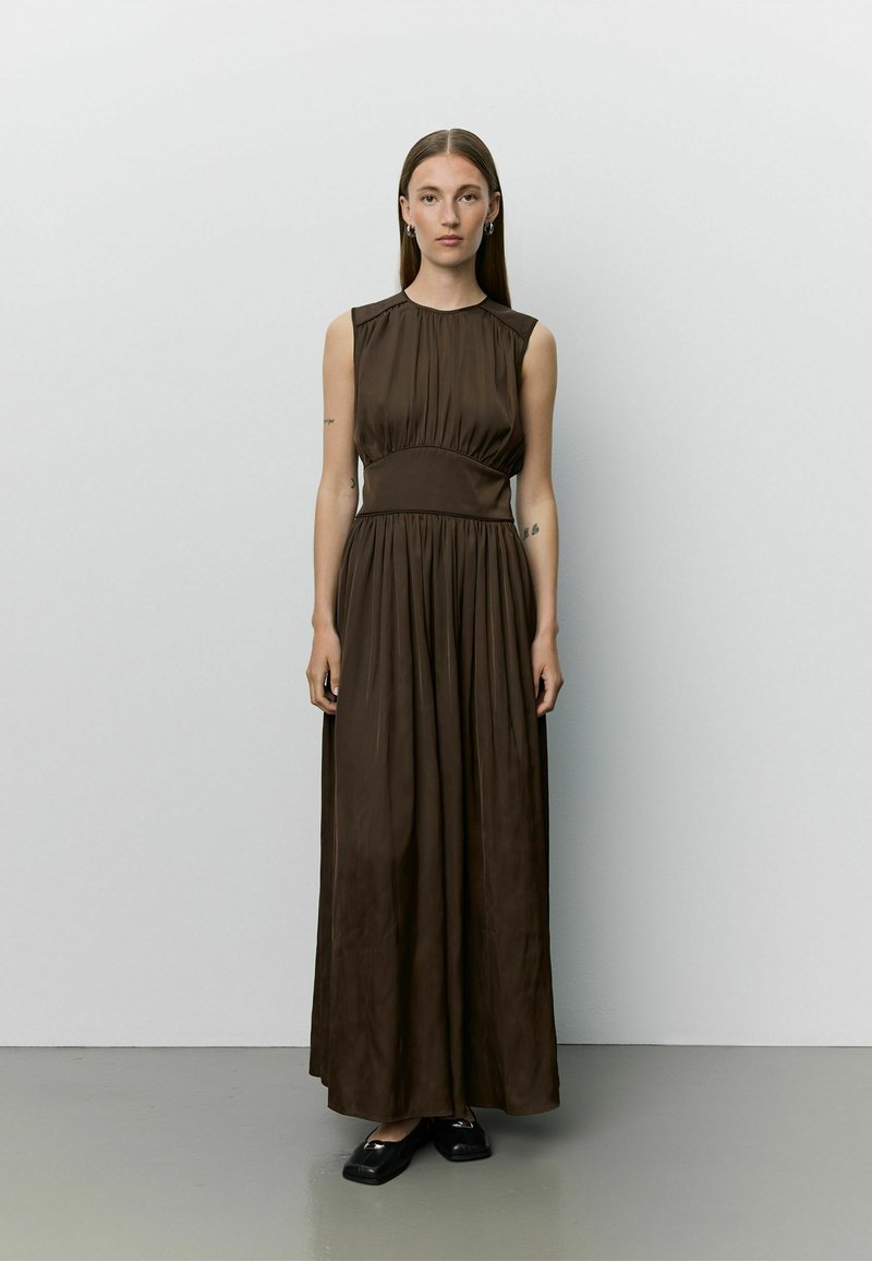 Femme aux longs cheveux lisses, portant une robe maxi marron foncé sans manches et des chaussures plates noires, debout contre un mur clair uni.
