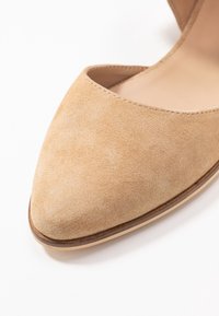 Beżowy zamszowy szpilka z wyciągniętym noskiem i płytkim przodem. Posiada gładką podeszwę i cienką, jasną krawędź. Minimalistyczny design.
