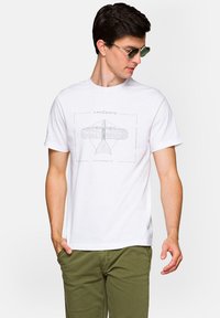 Biała bawełniana koszulka z grafiką vintage samolotu w odcieniu szarości. Krótkie rękawy, okrągły dekolt i luźny krój. Noszona z oliwkowymi spodniami.