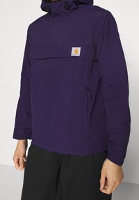 Lilla vandtæt jakke med hætte, frontlomme med klap og justerbare snore. Har elastiske manchetter og et logo-patch på brystet.