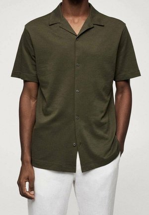 Homme portant une chemise boutonnée à manches courtes vert foncé et un pantalon blanc, une main dans la poche et l'autre détendue.