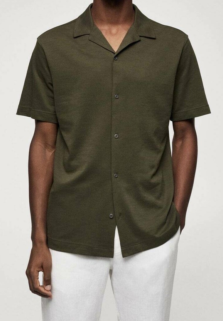 Homme portant une chemise boutonnée à manches courtes vert foncé et un pantalon blanc, une main dans la poche et l'autre détendue.