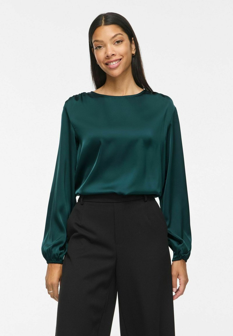 Femme aux longs cheveux foncés portant un chemisier en satin bleu sarcelle à manches longues et un pantalon noir taille haute, souriante sur un fond blanc uni.