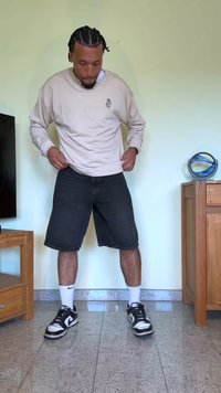 Camisa de manga longa bege clara, calções de denim pretos, sapatilhas Nike em preto e branco, meias brancas, de pé num chão liso com móveis de madeira por perto.