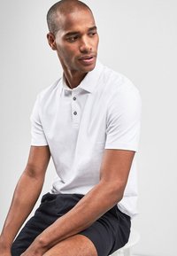 Hvid polo-shirt med en to-knaps placket og en klassisk krave, båret med navy shorts. Glat bomuldsstof, tætsiddende design, korte ærmer.