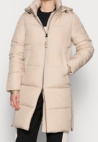 Abrigo puffer beige con capucha, cierre de cremallera, bolsillos laterales y diseño acolchado. Textura suave y longitud a media muslo con puños elásticos.