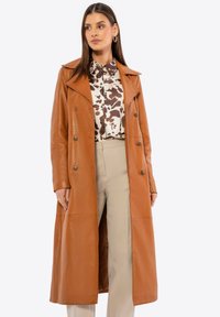 Abrigo trench de cuero marrón con un diseño de doble botonadura, cuello de muesca y textura suave, combinado con una blusa estampada y pantalones beige.