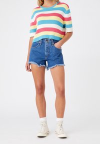 Woman wearing a short-sleeve rainbow-striped sweater, high-waisted frayed denim shorts, white socks, and white high-top sneakers.