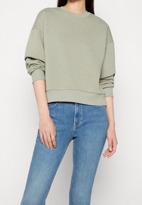 Mujer vistiendo un sudadera ligera en verde salvia con mangas largas y jeans ajustados de tiro alto en azul, frente a un fondo blanco liso.
