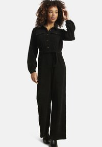 Tuta nera in velluto a coste con maniche lunghe, chiusura frontale con bottoni, due tasche sul petto, vita aderente e pantaloni a gamba larga. Decorata con bottoni marroni.