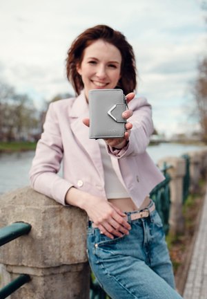 Young woman in pink jacket and jeans leans on riverside railing, smiling and holding a gray wallet toward the camera.