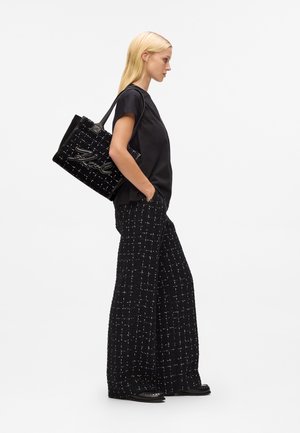 Sac fourre-tout en tissu tweed noir avec surface texturée et accents blancs, associé à un haut noir et à un pantalon large à motifs. Le mannequin pose de côté.