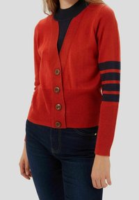 Red cardigan with brown buttons, featuring ribbed texture and navy stripes on sleeves. Paired with dark blue jeans.