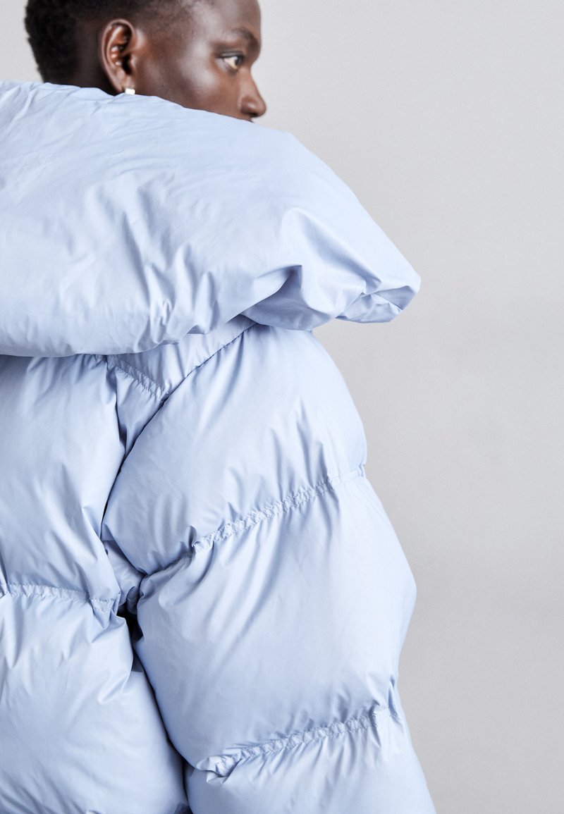 Doudoune bleu clair avec un col haut et une capuche surdimensionnée. Présente une texture matelassée et un ourlet élastiqué pour une allure ajustée.