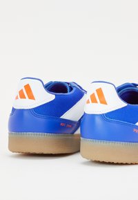 Blå sportssko med en overdel i mesh og syntetisk materiale, hvide detaljer, orange branding og en struktureret gummisål. Bagudsigten viser detaljer.