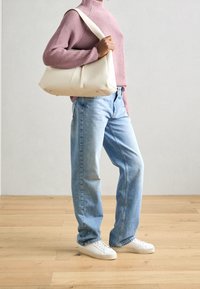Beige shoulder bag made of smooth material, paired with a pink ribbed turtleneck and blue relaxed jeans; model wears white sneakers.