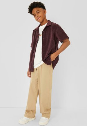 Teen boy standing with one hand in pocket, wearing a purple short-sleeve button-up shirt, white t-shirt, beige pants, and white sneakers.