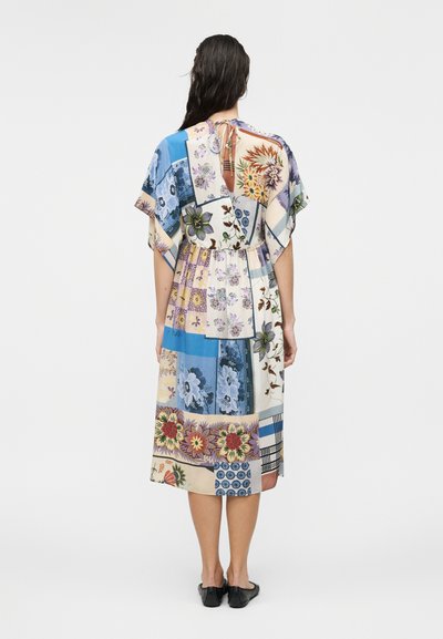 Femme aux longs cheveux foncés portant une robe midi fleurie multicolore patchwork avec des manches volantées, debout de dos à la caméra.