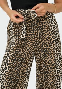 Pantalon à jambes larges à imprimé léopard, avec une ceinture nouée à la taille, une texture lisse et une palette de couleurs beige, noir et marron.