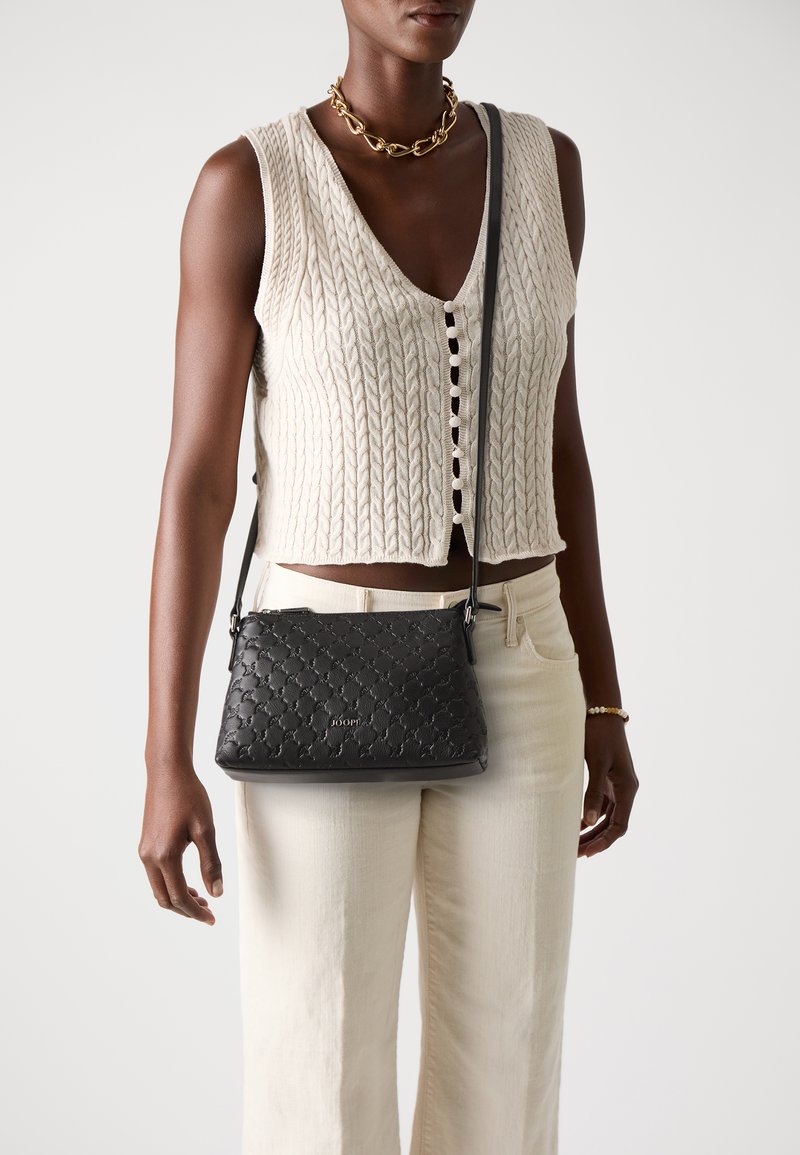 Una donna che indossa un top lavorato a maglia color crema e pantaloni chiari tiene in mano una borsa nera con texture, su uno sfondo semplice.