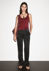Haut sans manches bordeaux, pantalon noir ajusté avec passants de ceinture et sandales plates. Le mannequin tient un sac texturé. Fond simple.