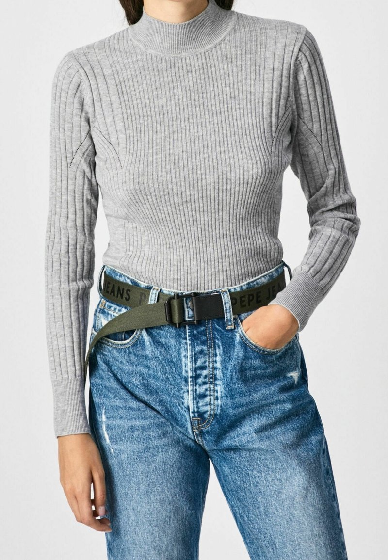 Femme portant un pull gris côtelé ajusté à col roulé, rentré dans un jean bleu taille haute avec une ceinture vert olive.