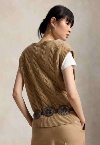 Kvinde med mørkt hår sat op i en knold har en beige kabelstrikket ærmeløs vest over en hvid kortærmet skjorte, beige bukser og et bælte med runde metalornamenter på.