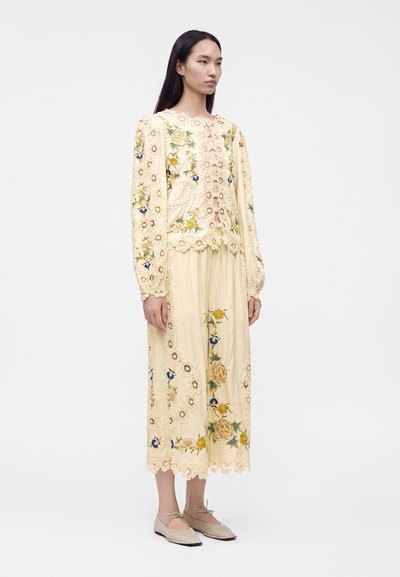 Femme portant un chemisier à manches longues crème brodé de fleurs et une jupe midi assortie avec des ballerines beiges, se tenant devant un fond blanc uni.