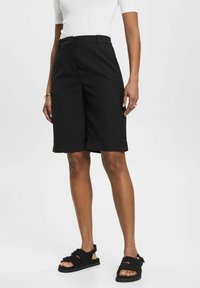 Shorts bermuda noirs à texture lisse, longueur genou, avec un pli avant et une ceinture simple. Associés à des sandales noires.