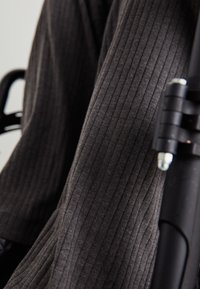 Um close-up de um tecido cinza escuro com texturas em relevo, proveniente de calças, parcialmente pendurado sobre o apoio de braço de uma cadeira de rodas negra, com hardware e detalhes de textura visíveis.