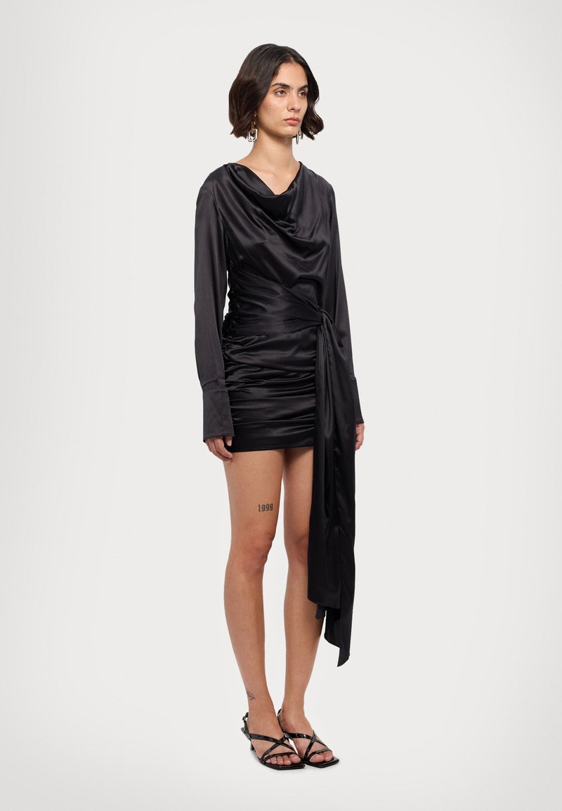 Vestido de satén negro con mangas largas, escote drapeado, silueta ajustada y detalle de lazo lateral. La modelo lleva sandalias negras de tiras.